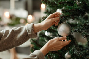 Hands reaching to hang a round, silver ornament on a Christmas tree