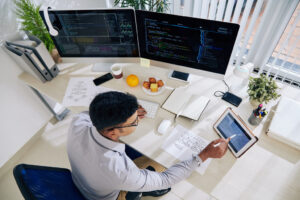 Software developer checks his calendar while working at a large, white desk with two monitors.